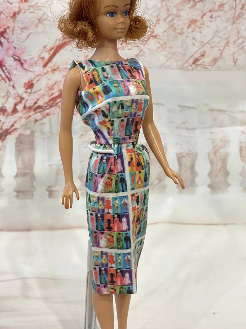 May include: A vintage doll wearing a colorful, patterned dress. The dress features a repeating design of figures in various outfits. The doll has red hair and blue eyes. The dress is sleeveless with a white belt.