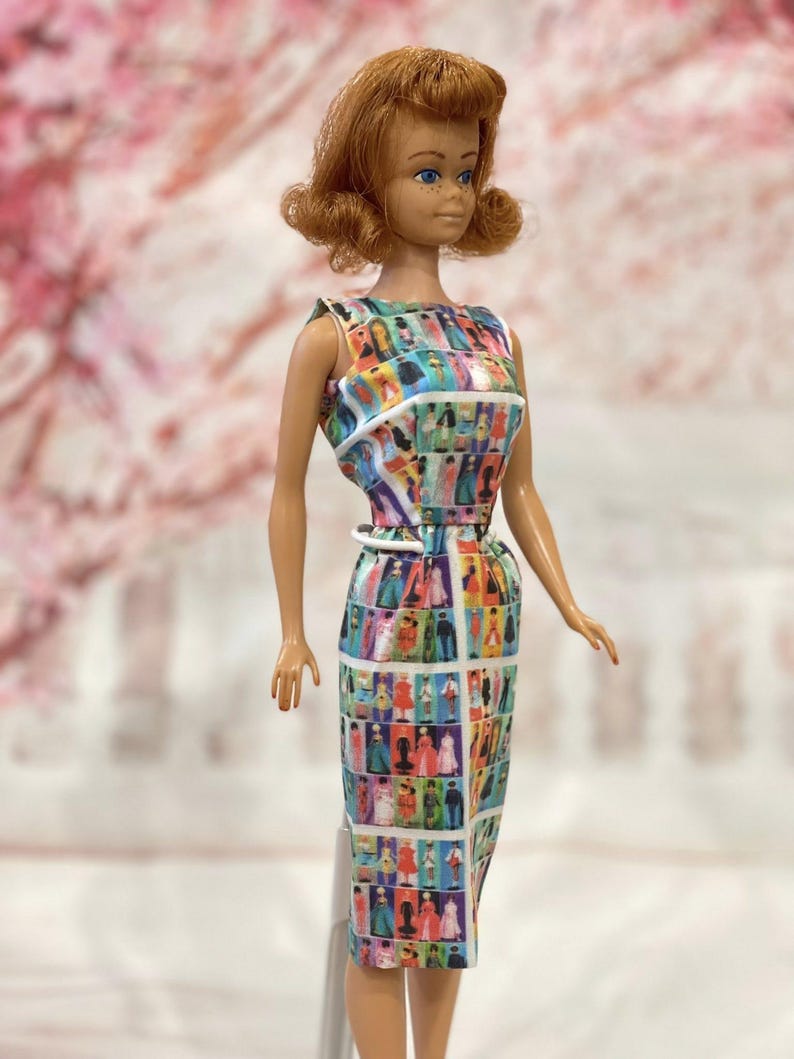 May include: A vintage doll with auburn hair and blue eyes, wearing a colorful sheath dress. The dress features a repeating pattern of figures in various outfits. The doll has a white belt and a white trim on the dress.