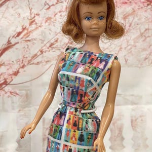 May include: A vintage doll with red hair and blue eyes, wearing a sleeveless dress. The dress features a colorful pattern of figures in various outfits, with a white belt. The doll is posed against a soft, floral background.