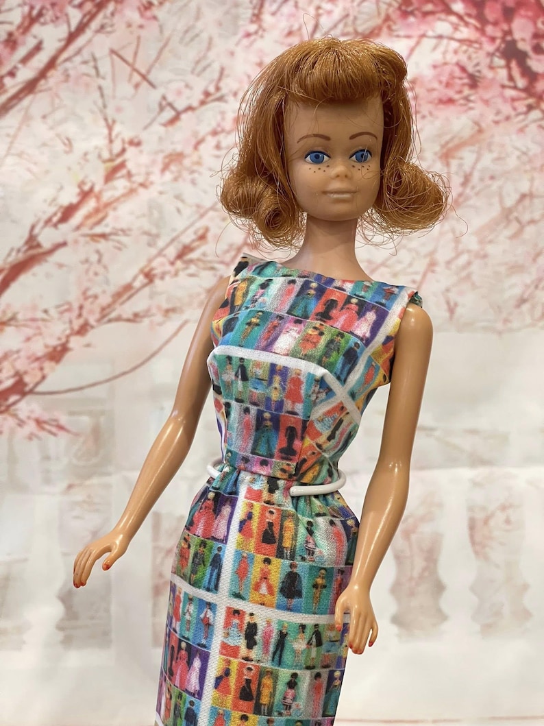 May include: A vintage doll with red hair and blue eyes, wearing a sleeveless dress. The dress features a colorful pattern of figures in various outfits, with a white belt. The doll is posed against a soft, floral background.