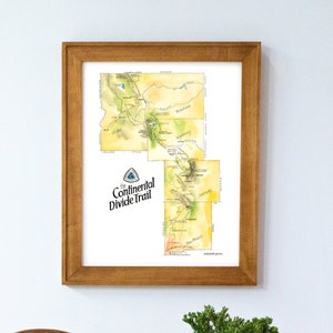 May include: A framed watercolor map of the Continental Divide Trail, with a wooden frame and a white mat. The map shows the trail's route through Montana, Wyoming, and Colorado. The map is hanging on a white wall, with a wooden basket and two potted plants in the foreground.