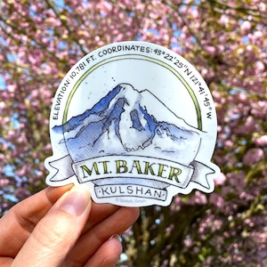 Pode incluir: Um adesivo branco com uma ilustração em aquarela do Monte Baker, uma montanha no estado de Washington. O adesivo apresenta a elevação da montanha, as coordenadas e o nome, "MT. BAKER", com a localização, "KULSHAN", escrita abaixo. O adesivo é segurado em uma mão contra um fundo de flores cor-de-rosa.