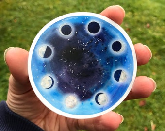 May include: A round sticker featuring a watercolour design of a blue night sky with white stars and a circle of eight different phases of the moon.