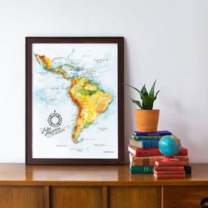 May include: A framed watercolor map of Latin America and the Caribbean. The map is in a brown frame and is hanging on a white wall. The map is surrounded by a stack of books and a potted plant.