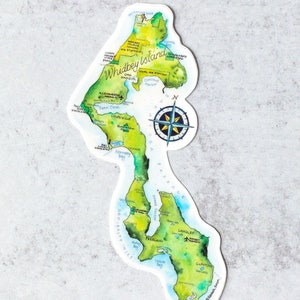 May include: A watercolour illustration of Whidbey Island, Washington, with a compass rose and key locations labelled. The island is depicted in shades of green, blue, and brown, with the water surrounding it in shades of blue.