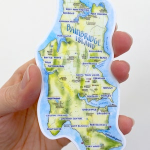 K&ouml;nnte beinhalten: Eine Aquarellkarte von Bainbridge Island, Washington, mit blauem Wasser und gr&uuml;nem Land. Die Karte zeigt die Lage verschiedener Sehensw&uuml;rdigkeiten, darunter Parks, Str&auml;nde und Stadtteile. Die Karte ist von einem wei&szlig;en Rand umgeben.
