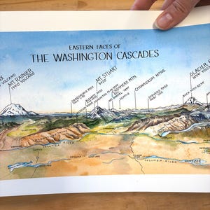 Może przedstawiać: Akwarelowa mapa zatytułowana "Eastern Faces of The Washington Cascades" przedstawiająca szczyty górskie, rzeki i szczegóły geograficzne. Zawiera nazwy takie jak Mt. Rainier, Mt. Stuart i Columbia River. Mapa jest trzymana przez dłoń.