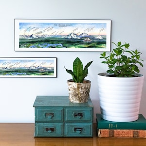 K&ouml;nnte beinhalten: Zwei gerahmte Illustrationen der Cascade Range im Bundesstaat Washington. Die obere Illustration ist gr&ouml;&szlig;er und zeigt die gesamte Kette, w&auml;hrend die untere Illustration kleiner ist und eine Nahaufnahme des Mount Rainier zeigt. Die Illustrationen h&auml;ngen an einer wei&szlig;en Wand &uuml;ber einer blaugr&uuml;nen Kommode mit vier Schubladen. Eine Topfpflanze und eine Topfminze stehen auf der Kommode, zusammen mit einem Stapel B&uuml;cher.