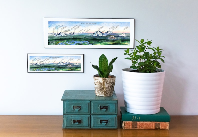 K&ouml;nnte beinhalten: Zwei gerahmte Illustrationen der Cascade Range im Bundesstaat Washington. Die obere Illustration ist gr&ouml;&szlig;er und zeigt die gesamte Kette, w&auml;hrend die untere Illustration kleiner ist und eine Nahaufnahme des Mount Rainier zeigt. Die Illustrationen h&auml;ngen an einer wei&szlig;en Wand &uuml;ber einer blaugr&uuml;nen Kommode mit vier Schubladen. Eine Topfpflanze und eine Topfminze stehen auf der Kommode, zusammen mit einem Stapel B&uuml;cher.