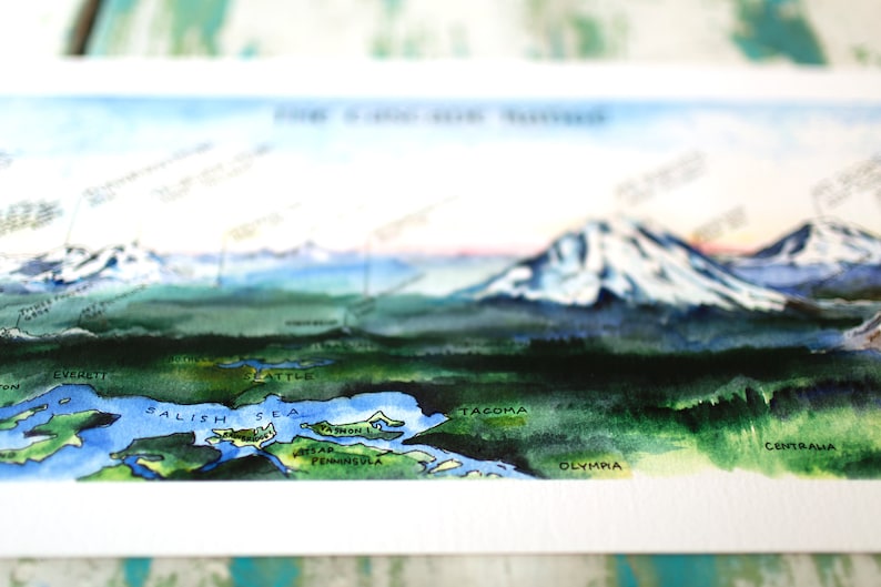 K&ouml;nnte beinhalten: Aquarellmalerei einer Landschaft mit Bergen, einem See und dem Text "Salish Sea" in Blau. Die Berge sind schneebedeckt und der See ist von gr&uuml;nen B&auml;umen umgeben. Der Text "Everett", "Seattle", "Tacoma" und "Olympia" ist ebenfalls auf dem Bild zu sehen.