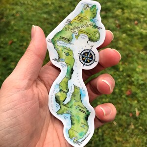 May include: A sticker of Whidbey Island, Washington, with a watercolour-style map of the island. The sticker is white with a green and blue map of the island, and includes the names of towns and points of interest. The sticker also includes a compass rose.