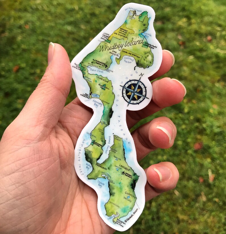 May include: A sticker of Whidbey Island, Washington, with a watercolour-style map of the island. The sticker is white with a green and blue map of the island, and includes the names of towns and points of interest. The sticker also includes a compass rose.