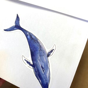 May include: A watercolor sticker of a blue whale. The whale is depicted in shades of blue, with a white outline. The sticker is on a white background. The whale's tail and fins are visible.