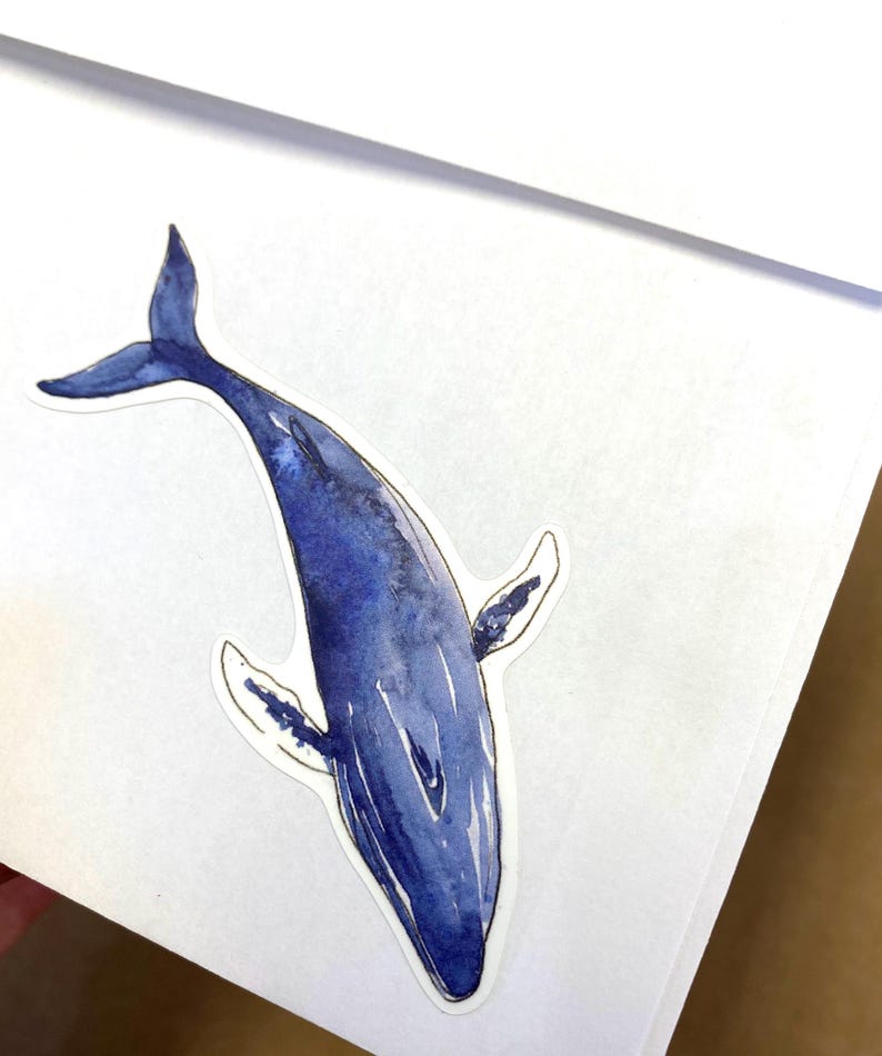 May include: A watercolor sticker of a blue whale. The whale is depicted in shades of blue, with a white outline. The sticker is on a white background. The whale's tail and fins are visible.