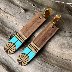 May include: A pair of wooden earrings with a geometric design. The earrings are painted in shades of blue and gold, and feature a sun-like design. The earrings are attached to gold-coloured hooks.