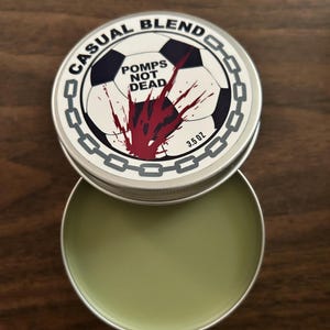 Op de afbeelding: Een ronde metalen blik met lichtgroene balsem, met een deksel voorzien van een voetbalafbeelding, een rood verfspat-effect, een kettingrand en de tekst CASUAL BLEND en POMPS NOT DEAD, met een nettogewicht van 3,5 oz op het etiket.