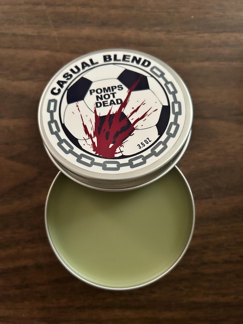 Op de afbeelding: Een ronde metalen blik met lichtgroene balsem, met een deksel voorzien van een voetbalafbeelding, een rood verfspat-effect, een kettingrand en de tekst CASUAL BLEND en POMPS NOT DEAD, met een nettogewicht van 3,5 oz op het etiket.