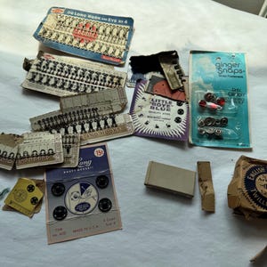 May include: A collection of vintage sewing notions including De Long hook and eye sets, Ginger Snaps fasteners, Little Boye Blue quality steel fasteners, and various metal buttons on cardboard backing cards with printed text and branding.
