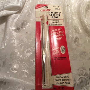 May include: A silver crochet hook with a red and white label. The label reads "Susan Bates Clipson "Steelite" Crochet Hook" and "Exclusive Time-Saver Stitch Saver Clip". The label also says "Crochet with Ease-Other Sister" and "Exclusive micro-ground® in-line® head No. 12205".
