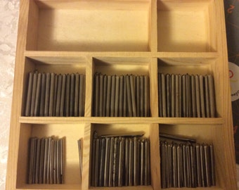 May include: A wooden box with multiple compartments filled with small, silver-colored metal rods. The box is made of light wood and has a simple design.