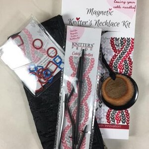 May include: A black zippered pouch with a magnetic knitter's necklace kit. The kit includes two black cable needles, a black and brown wooden bead, and a white and red patterned fabric with the text "Knitters Pride" and "with Innovation!"
