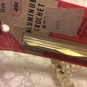Pode incluir: Um gancho de crochê de alumínio dourado com o texto "MADE C USA" na alça. O gancho está embalado em um cartão rosa e branco com o texto "ALUMINUM CROCHET HOOK" e "49¢".