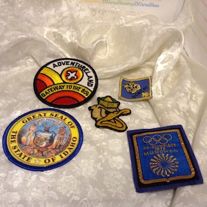 Pode incluir: Uma coleção de seis patches bordados. Os patches são de várias formas e tamanhos e apresentam diferentes designs. Um patch é amarelo e vermelho com o texto "Adventureland Gateway to the 80s". Outro patch é azul e amarelo com o texto "Bobcat". Um terceiro patch é azul e dourado com o texto "XX. Olympiade 1972 Munchen". Um quarto patch é azul e amarelo com o texto "The State of Idaho". Um quinto patch é amarelo e verde com o texto "Oregon". O sexto patch é amarelo e vermelho com um design de pato.