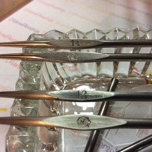 Peut inclure: Gros plan sur des crochets en métal vintage. Deux crochets affichent le numéro 14 et deux le numéro 13. Les crochets sont de couleur argent et bronze, avec un emblème décoratif. Les crochets reposent dans un plat en verre transparent.