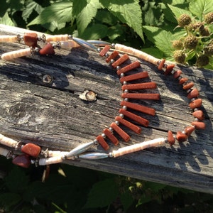 Könnte beinhalten: Eine handgefertigte Halskette mit abwechselnden Abschnitten aus cremefarbenen, geriffelten Perlen und rotbraunen, rechteckigen und unregelmäßig geformten Steinen. Die Halskette hat silberfarbene Metallakzente und wird auf einer verwitterten Holzoberfläche präsentiert.