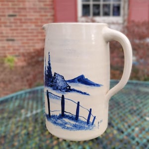 May include: A white ceramic pitcher with a large handle. The pitcher is decorated with a blue landscape scene featuring a fence, mountains, and trees. The pitcher is sitting on a green metal table.