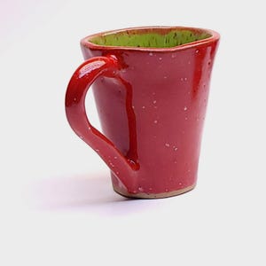 May include: A handmade red ceramic mug with a green interior. The mug has a rounded handle and a speckled finish. The mug is approximately 4 inches tall.