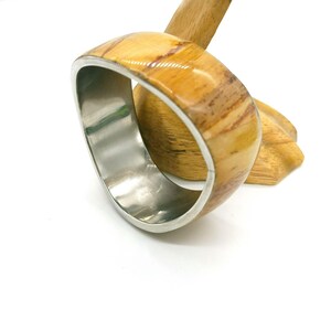 May include: A wide, oval-shaped bracelet with a polished wood exterior and a silver-colored metal interior. The wood grain features shades of tan and brown. The bracelet is displayed on a wooden stand against a white background.