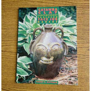 May include: A softcover book titled Catawba Clay: Contemporary Southern Face Jug Makers by Barry G. Huffman. The cover features a dark brown, glazed ceramic face jug with two handles and a sculpted human face, set against green foliage.