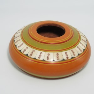 May include: A round, orange decorative bowl with a central opening. The bowl features a green and red band around the top, and is decorated with white, bone-shaped accents. The bowl is likely handmade and has a rustic aesthetic.