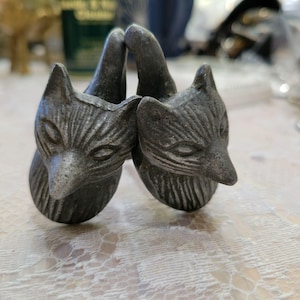 May include: Two antique silver-colored fox head napkin rings. The fox heads are facing each other, with detailed facial features and pointed ears. The napkin rings are made of metal and have a vintage aesthetic.