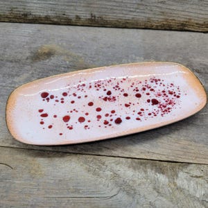 May include: A rectangular serving dish with a white interior speckled with red dots. The dish has a light pink border and a dark brown edge. The dish is placed on a weathered wooden surface.