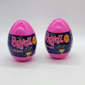 May include: Two pink egg-shaped containers with the word "Bratz" in pink lettering. The containers have a dark blue band with the word "Stickers" and a cartoon image of a Bratz doll. The containers are on a white surface.