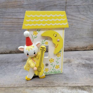 May include: A vintage, whimsical light switch cover featuring a cartoon character with a red hat and yellow guitar. The cover is a white house with a yellow roof and moon, decorated with floral accents. The overall design is charming and nostalgic.