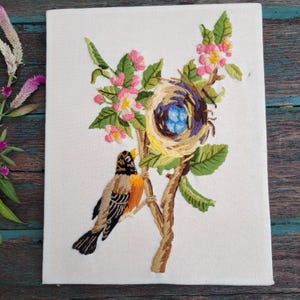 May include: A rectangular canvas featuring a detailed embroidery of a bird perched on a branch next to a nest containing three blue eggs. The design includes green leaves and pink flowers, with the bird displaying brown, black, and orange plumage.
