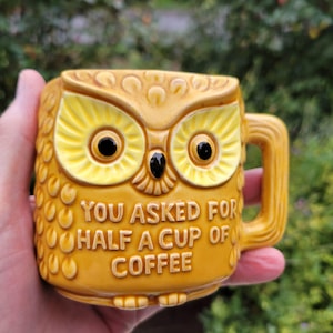 May include: A yellow ceramic owl-shaped mug with a textured feather pattern and a handle. The front features the embossed text "YOU ASKED FOR HALF A CUP OF COFFEE" in capital letters. The owl has large yellow eyes with black pupils.