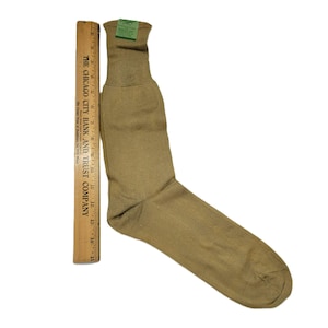 May include: A single tan-colored sock with a ribbed cuff and a tag attached. A wooden ruler is placed next to the sock for scale. The sock appears to be made of a soft, possibly wool, material.