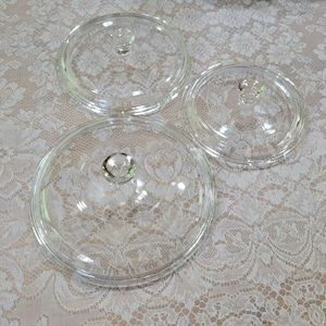 May include: Three clear glass pot lids with round knobs. The lids are of varying sizes and are made of transparent glass. The knobs are also clear and round. The lids are arranged on a white lace tablecloth.