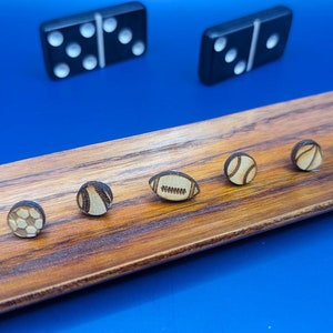 May include: A wooden tray with six small wooden circles, each with a different sports ball design. The designs include a baseball, basketball, football, soccer ball, golf ball, and a rugby ball. The tray is on a blue surface.