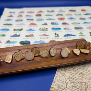 May include: Wooden pins and earrings with travel and map themes. Includes Route 66, National Park, and State Park designs. The earrings have dates and map details. The items are displayed on a wooden board with a map background.