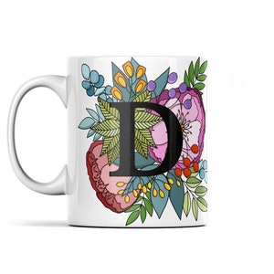 Può includere: Tazza in ceramica bianca con una grande lettera "D" nera circondata da un colorato motivo floreale. I fiori includono fiori rosa, rossi, blu e gialli con foglie verdi. La tazza ha un manico curvo.