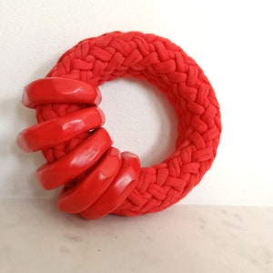 Könnte beinhalten: Eine leuchtend rote Halskette mit einem kreisförmigen, geflochtenen Design. Sechs glänzende, rote Holzringe sind auf einer Seite gestapelt. Der Schmuck ist ein auffälliges Accessoire.