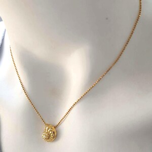 May include: A gold-colored necklace with a small, textured pendant. The pendant has a detailed, possibly floral or shell-like design. The chain is delicate and appears to be made of small beads. The necklace is displayed on a mannequin.