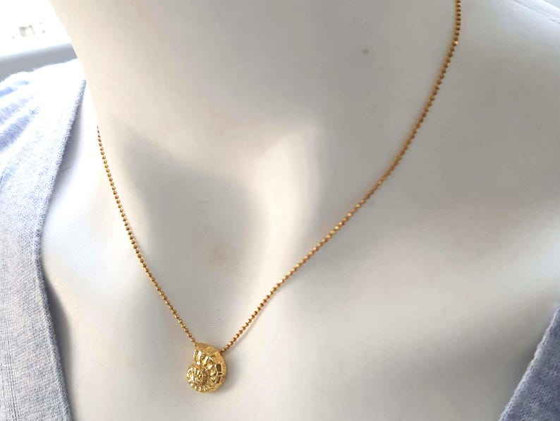 May include: A gold-colored necklace with a small, textured pendant. The pendant has a detailed, possibly floral or shell-like design. The chain is delicate and appears to be made of small beads. The necklace is displayed on a mannequin.