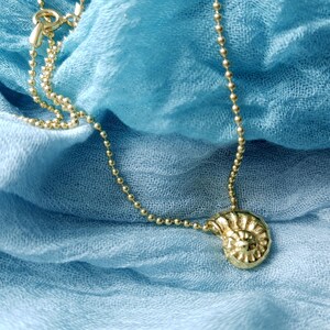 May include: A gold-colored necklace with a pendant shaped like a spiral seashell. The necklace chain is made of small, round beads. The jewelry rests on a blue fabric background.