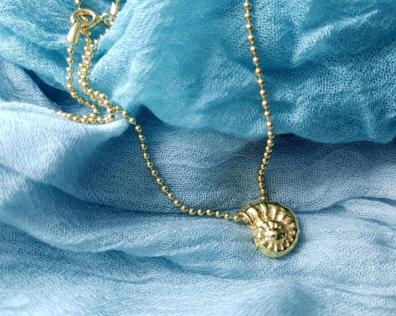 May include: A gold-colored necklace with a pendant shaped like a spiral seashell. The necklace chain is made of small, round beads. The jewelry rests on a blue fabric background.
